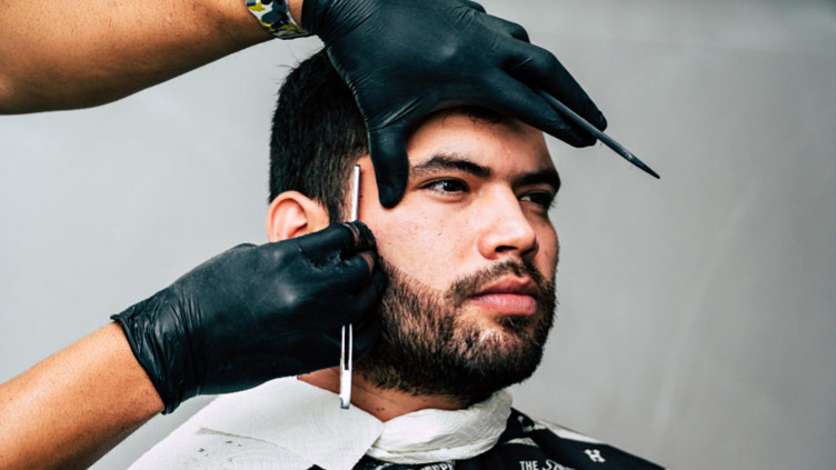 A young man is having his beard shaven by a barber in gloves