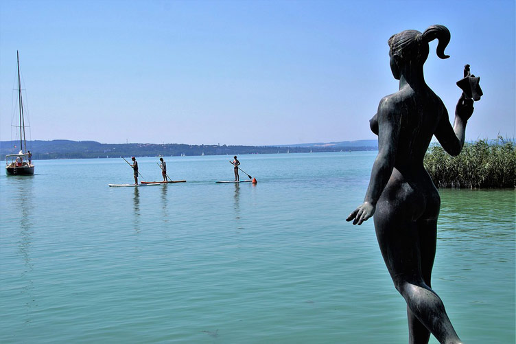 Statue of a female facing the Balaton lake, and pepole enjoying water activities in the lake