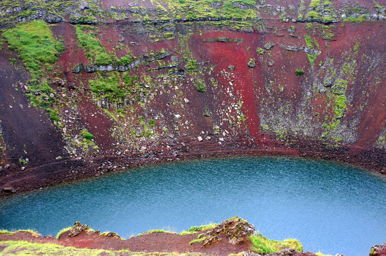 Blue water of Kerid crater volcanic lake surrounded by red slopes covered in patches of green vegetation