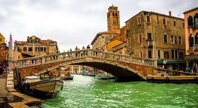 A bridge over the green waters of the Grand Canal of Venice, boats and historical building in the background