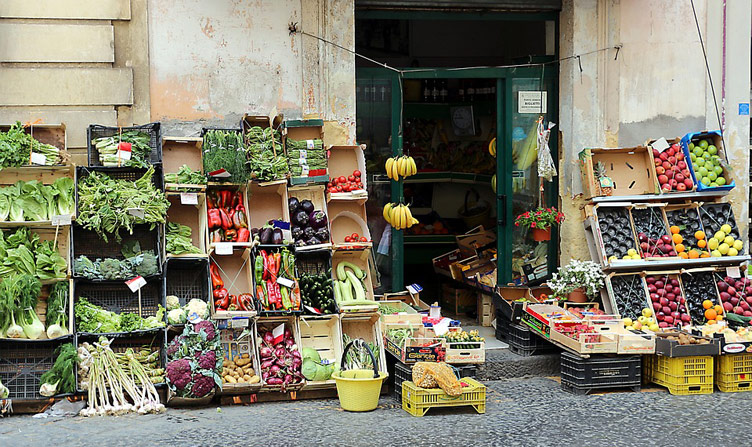 The front of a very basic fruit and veg shop on a sidestreet in Sicily. The shop has fruit and veg stalls and boxes on display. 