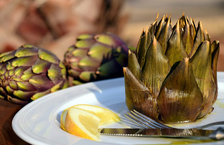 Ready to eat artichoke served on a plate with a slice of lemon.