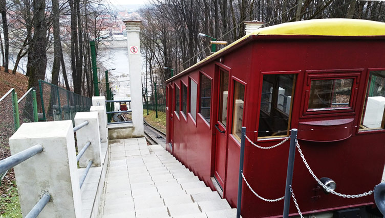 A dark red vintage funicular tram on top of a hill with a winter backdrop of trees and views of a river and city rooftops