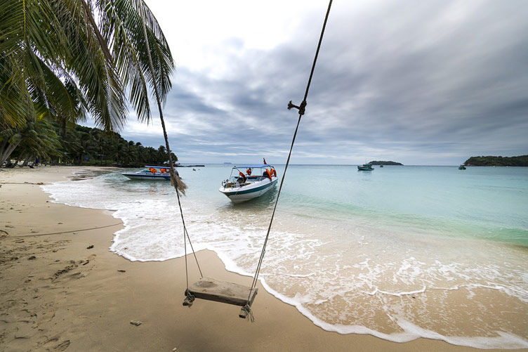 A swing, a boat, a palm tree set against a backdrop of Phu Quoc white sand beach and crystal clear blue ocean waters 